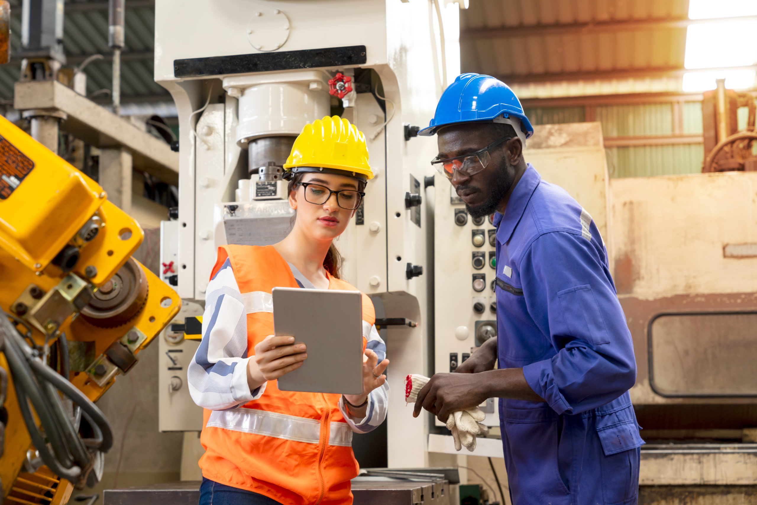 woman wearing hard hat consults with man wearing hard hat while looking at a tablet in a manufacturing facility